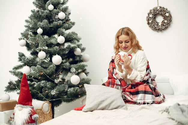 Vrouw in een kamer. Blondje in een witte trui. Dame dichtbij kerstboom.