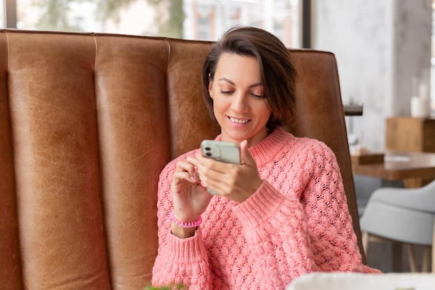 Vrouw in een restaurant in een warme trui kijkt naar iets aan de telefoon