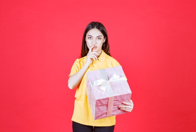 vrouw in geel shirt met een roze geschenkdoos en ziet er verward en aarzelend uit.