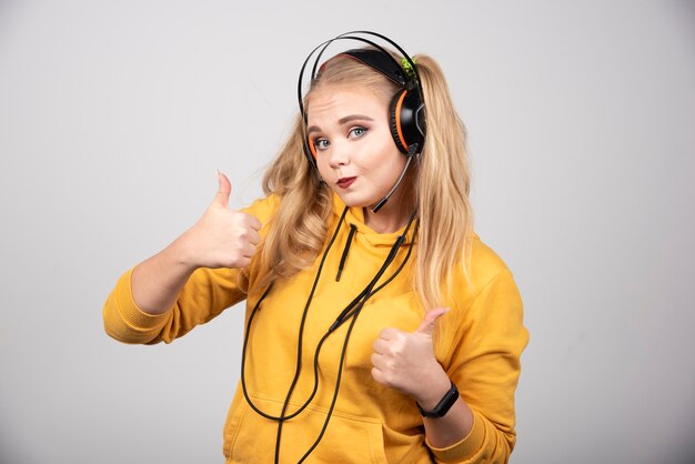 Vrouw in geel sweatshirt die duimen op grijze achtergrond opgeven.