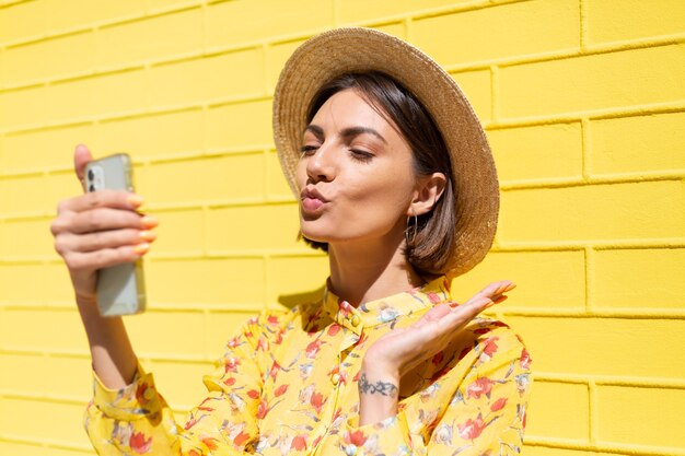 Vrouw in gele zomerjurk en hoed op gele bakstenen muur kalm en positief bedrijf mobiele telefoon