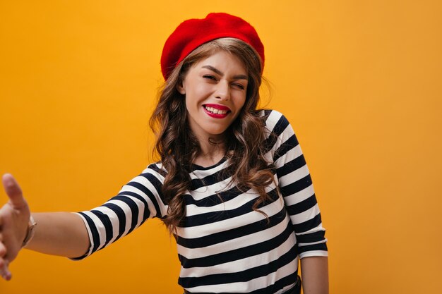 Vrouw in gestreept overhemd en baret knipogen en selfie nemen op oranje achtergrond. Aantrekkelijk meisje in modieuze outfit poseren.