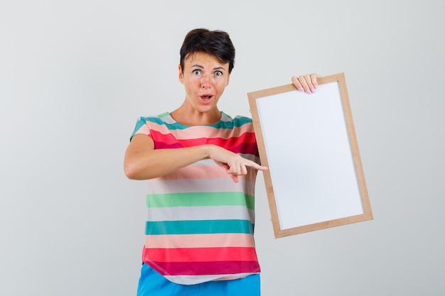 Vrouw in gestreept t-shirt, broek wijzend op leeg frame en op zoek verrast, vooraanzicht.