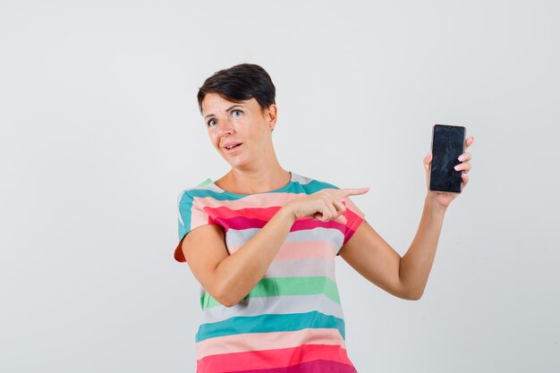 Vrouw in gestreept t-shirt wijzend op mobiele telefoon en aarzelend, vooraanzicht op zoek.