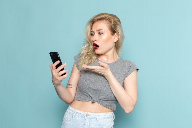 vrouw in grijs shirt en heldere blauwe spijkerbroek met een telefoon met verbaasde uitdrukking