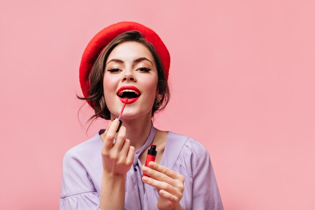 Vrouw in heldere baret schildert haar lippen met rode lippenstift. Meisje in lila blouse poseren op roze achtergrond.