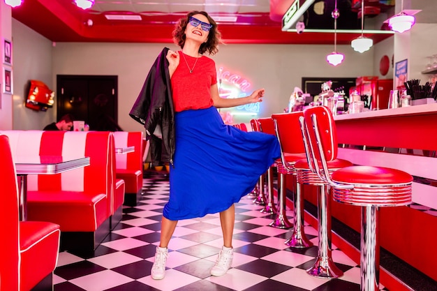 vrouw in kleurrijke outfit in retro vintage jaren 50 café dansen met jas, blauwe rok en rood shirt, zonnebril met plezier in vrolijke stemming