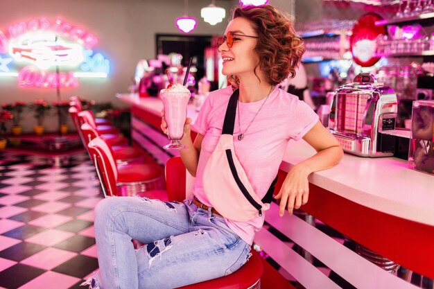 vrouw in kleurrijke retro vintage amerikaanse jaren 50 café zittend aan tafel en drink milkshake cocktail met roze zonnebril, plezier