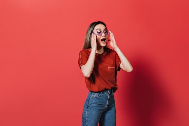 Vrouw in lila bril en rood T-shirt emotioneel poseren op geïsoleerde muur