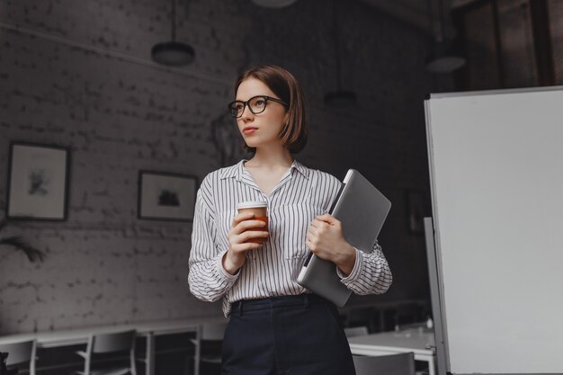 Vrouw in office broek en shirt vormt met kopje koffie en laptop bezit. Schot van kortharig meisje in glazen in heldere kantoor.