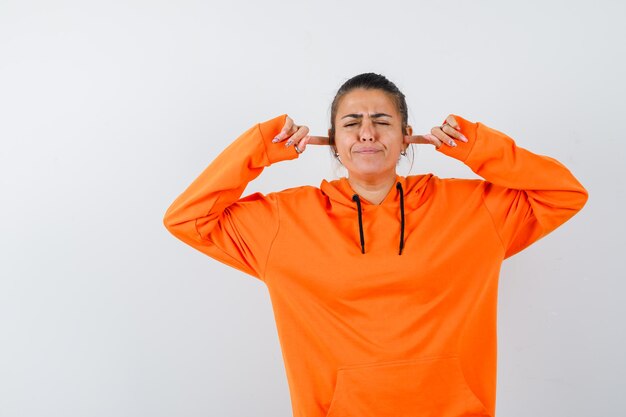 Vrouw in oranje hoodie die oren dichtstopt met vingers en er geïrriteerd uitziet