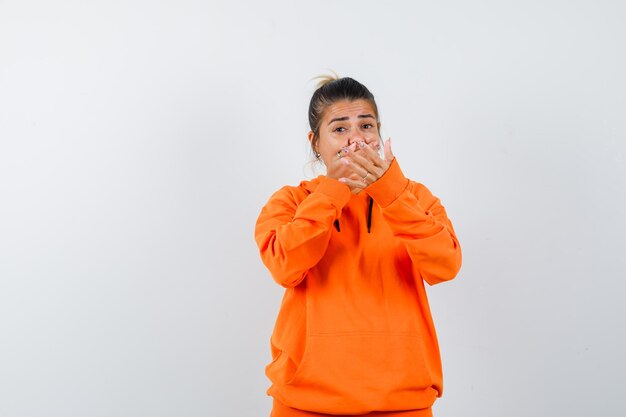 vrouw in oranje hoodie die stopgebaar toont, hand op mond houdt en bang kijkt