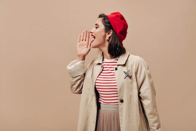 Vrouw in rode baret en loopgraaf schreeuwen op beige achtergrond. Stijlvol meisje met heldere lippen in gestreepte trui en modieuze jas vormt voor camera.