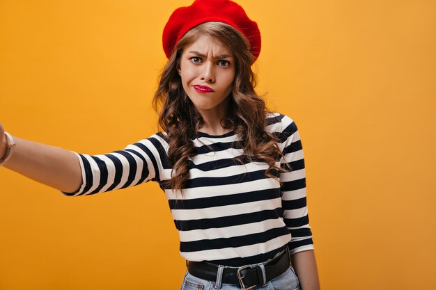 Vrouw in rode baret kijkt bedachtzaam en maakt selfie Stijlvol jong meisje in gestreepte blouse en heldere baret neemt foto
