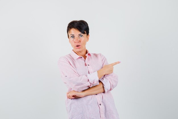 Vrouw in roze shirt wijst naar de rechterkant en kijkt verbaasd, vooraanzicht.