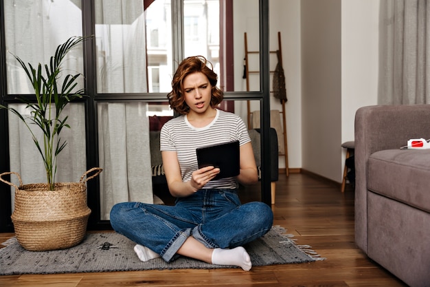 Vrouw in spijkerbroek zit op de vloer in appartement en kijkt met misverstand naar haar tablet.