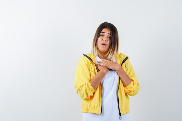 Vrouw in t-shirt, jas die handen op de borst houdt en verbaasd, vooraanzicht kijkt.