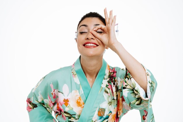 Vrouw in traditionele Japanse kimono die ok teken doet die door vingers kijkt gelukkig en vreugdevol glimlachend op wit