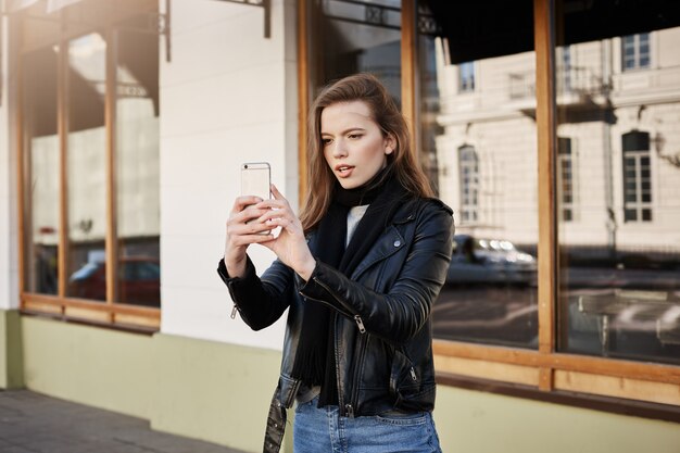 vrouw in trendy leren jas met smartphone terwijl ze een foto maakt van een landschap of band die op straat speelt