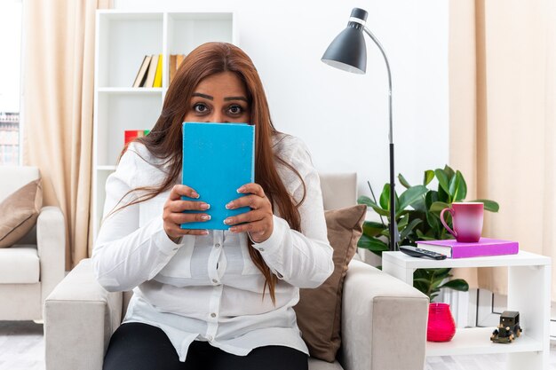 Vrouw in wit overhemd en zwart met boek dat gezicht bedekt met boek bezorgd zittend op de stoel in lichte woonkamer