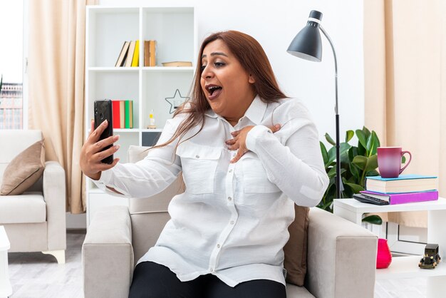 Vrouw in wit overhemd en zwarte broek doet selfie met smartphone gelukkig en zelfvoldaan zittend op de stoel in lichte woonkamer