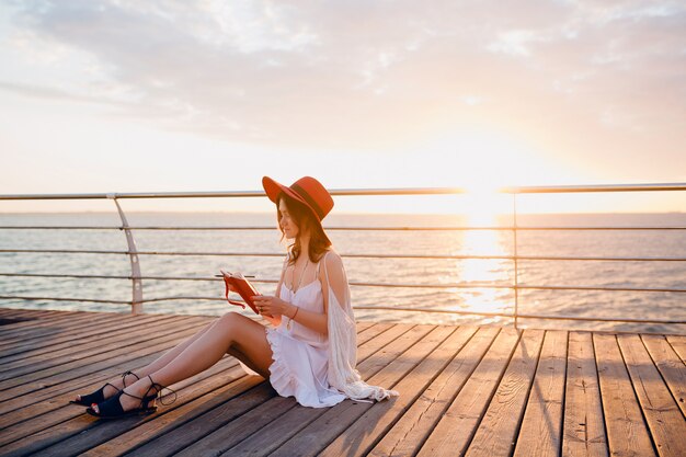 vrouw in witte jurk zittend aan zee op zonsopgang denken en het maken van aantekeningen in dagboek boek in romantische stemming met rode hoed