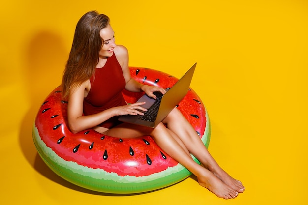 Vrouw in zwembroek zit in opblaasbare ring werkt op laptop op gele achtergrond freelance reizen
