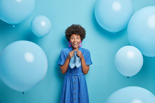 vrouw jurken voor speciale gelegenheid draagt jurk houdt schoenen met hoge hakken heeft vrolijke uitdrukking poses rond opgeblazen ballonnen geïsoleerd op blauw