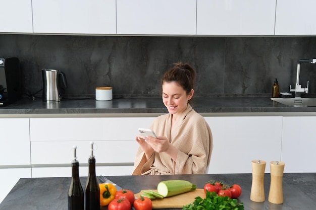 Vrouw kijken naar recept op smartphone zittend in de keuken met groenten en snijplank