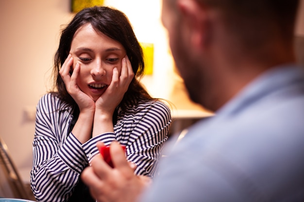 Vrouw kijkt blij naar verlovingsring tijdens huwelijksaanzoek terwijl ze een romantisch diner heeft. Man vraagt zijn vriendin ten huwelijk in de keuken tijdens een romantisch diner. Gelukkige blanke vrouw die lacht be