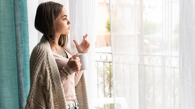 Vrouw kijkt door het raam thuis tijdens de pandemie terwijl ze koffie drinkt