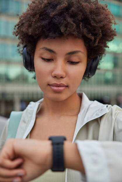 vrouw kijkt naar smartwatch op pols volgt gezondheid na training luistert favoriete audiotrack in koptelefoon gekleed in jashoudingen