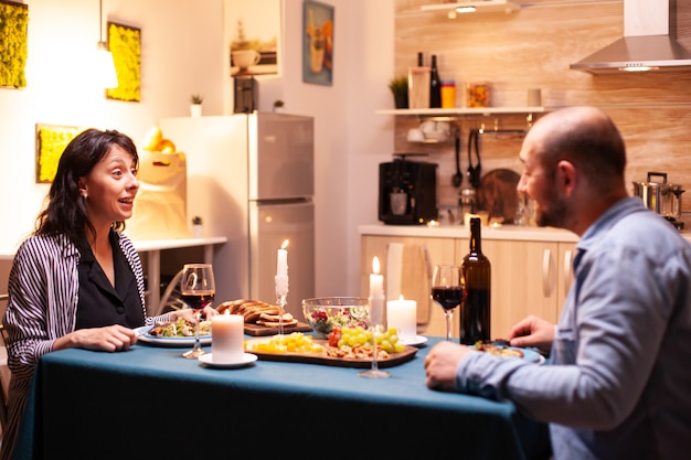 Vrouw kijkt verbaasd naar man tijdens romantisch diner in de keuken. Praten gelukkig zittend aan tafel eetkamer, genietend van de maaltijd thuis met romantische tijd bij kaarslicht.