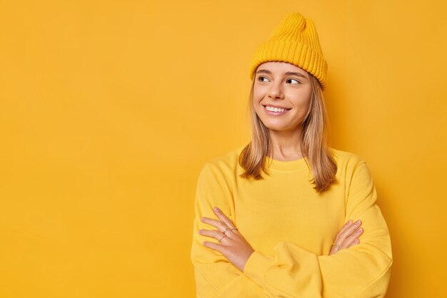 vrouw kijkt weg met blije uitdrukking houdt de armen gevouwen heeft een doordachte blik draagt een casual trui en hoed geïsoleerd op fel geel met kopieerruimte