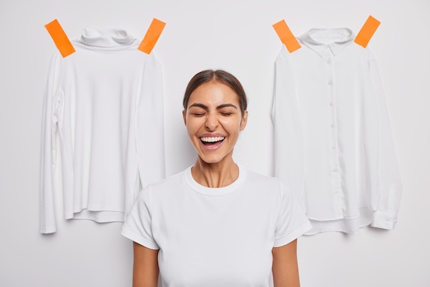 Gratis foto vrouw lacht vrolijk houdt ogen gesloten, gekleed in casual t-shirt poses op wit met gepleisterde coltrui en shirt gaan zich kleden voor straatwandeling. mensen en kleding