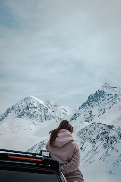 Vrouw leunend op een zwart voertuig voor verbazingwekkende besneeuwde en rotsachtige bergen en bewolkte luchten