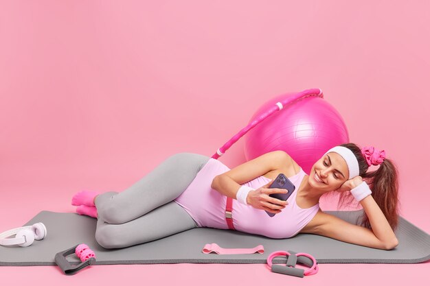 vrouw ligt op comfortabele fitnessmat kijkt naar video via smartphone gekleed in sportkleding oefeningen met sportuitrusting