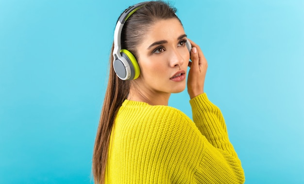 vrouw luisteren naar muziek in draadloze koptelefoon gelukkig dragen gele gebreide trui poseren op blauw