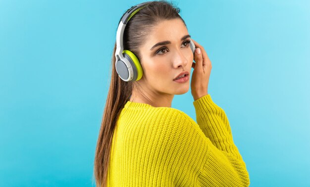 vrouw luisteren naar muziek in draadloze koptelefoon gelukkig dragen gele gebreide trui poseren op blauw
