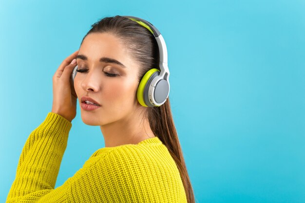vrouw luisteren naar muziek in draadloze koptelefoon gelukkig dragen gele gebreide trui poseren op blauw