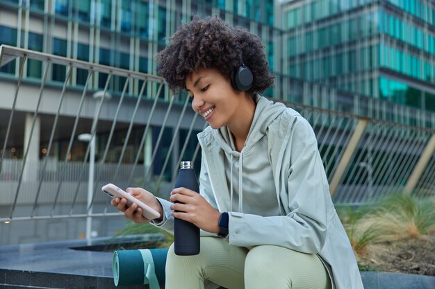 vrouw luistert muziek in koptelefoon scrollt sociale netwerken via smartphone neemt pauze na training poseert buiten tegen modern glazen gebouw in goed humeur