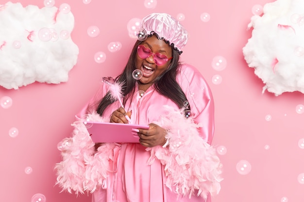 vrouw maakt aantekeningen in dagboek geniet van huiselijke sfeer draagt badmuts kamerjas zonnebril lacht vrolijk poseert op roze