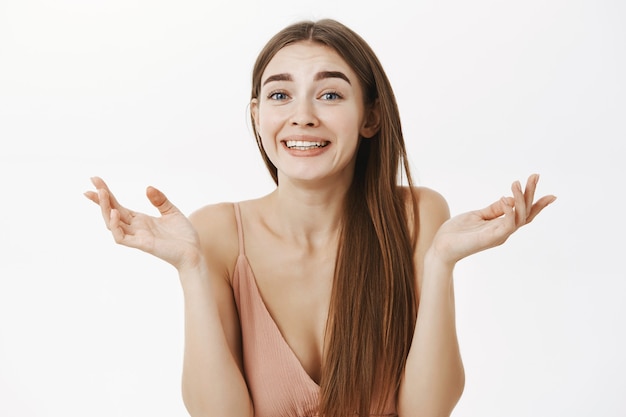 Vrouw maakt oprechte verbaasde uitdrukking, schouderophalend met handen uitgespreid in de buurt van schouders glimlachend en verbaasd staande onder de indruk en verbaasd in beige jurk