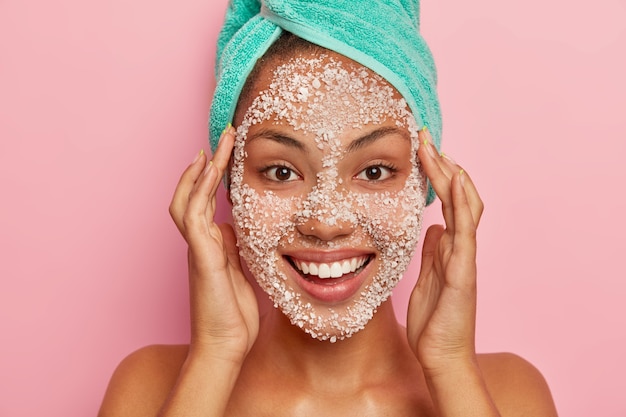 Vrouw masseert gezicht, past een natuurlijk scrubmasker toe, reinigt de gezichtshuid voor een jonge uitstraling, heeft manicure, draagt een gewikkelde handdoek op het hoofd, geïsoleerd op een roze muur