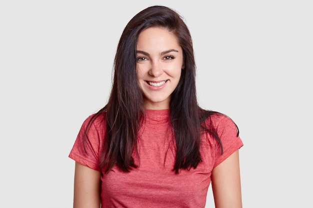 Vrouw met aangename glimlach, donker haar, gekleed in casual roze t-shirt, heeft witte perfecte tanden, verheugt zich over het ontvangen van complimenten