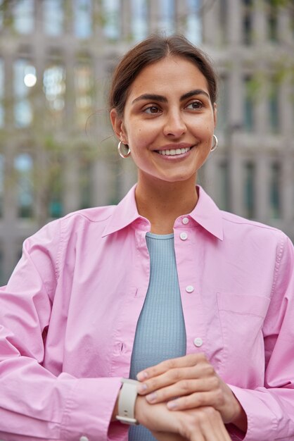 vrouw met aangename uitstraling wacht op iemand heeft afspraak controleert tijd op horloge glimlacht zachtjes staat buiten op wazig architectonisch gebouw draagt roze shirt.