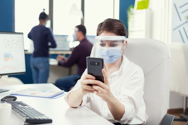 Vrouw met beschermend masker die in een professionele werkruimte aan het typen op een mobiele telefoon achter de computer werkt