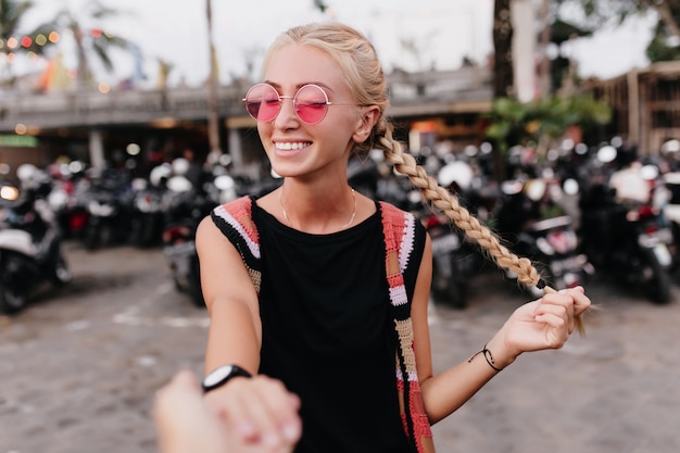 vrouw met blij gezicht expressie poseren in trendy roze zonnebril. speels gelooid vrouwelijk model haar vlechten aan te raken en te lachen.