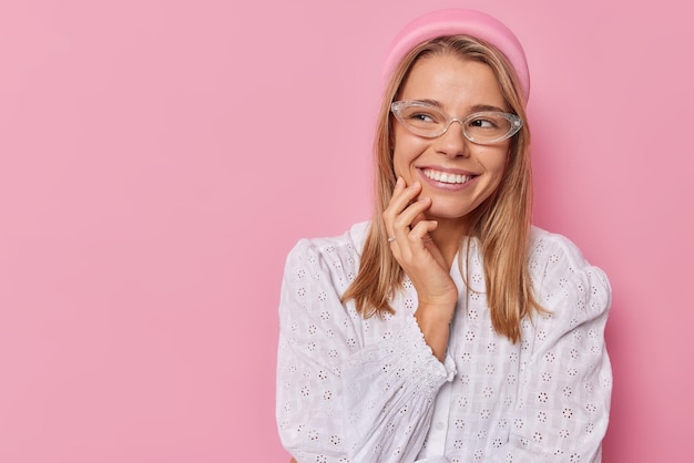 Vrouw met blije uitdrukking houdt hand op gezicht hoort aangenaam nieuws draagt transparante bril witte blouse geconcentreerd weg geïsoleerd op roze blanco kopieerruimte