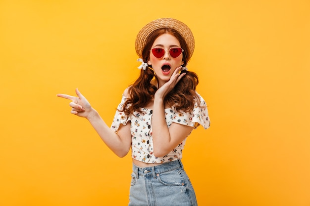 Vrouw met bogen op haar haar en hoed draagt camera kijkt verbaasd. vrouw in rode zonnebril en denim rok wijst vinger naar geïsoleerde achtergrond.